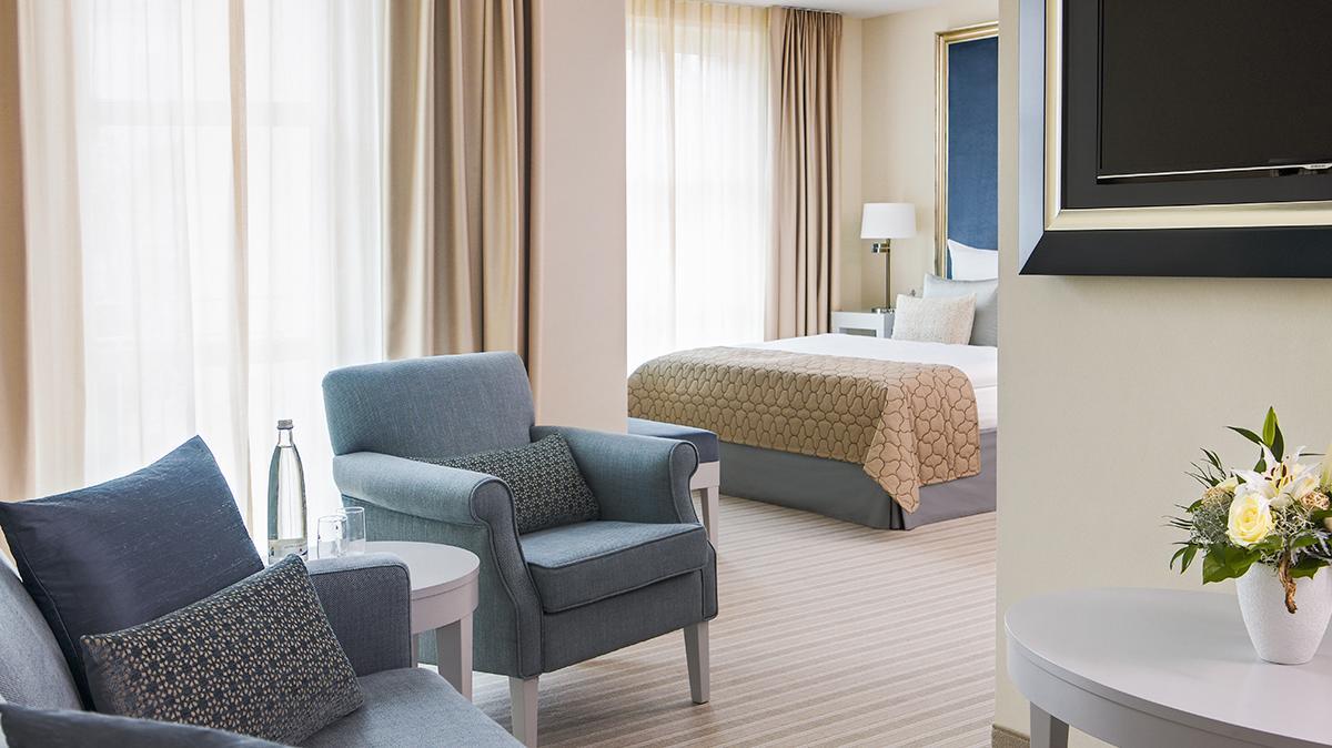 Sitzgruppe aus blauem Stoff in einem eleganten Hotelzimmer mit langen transparent-beigen Vorhängen und Bett mit beiger Husse im Hintergrund